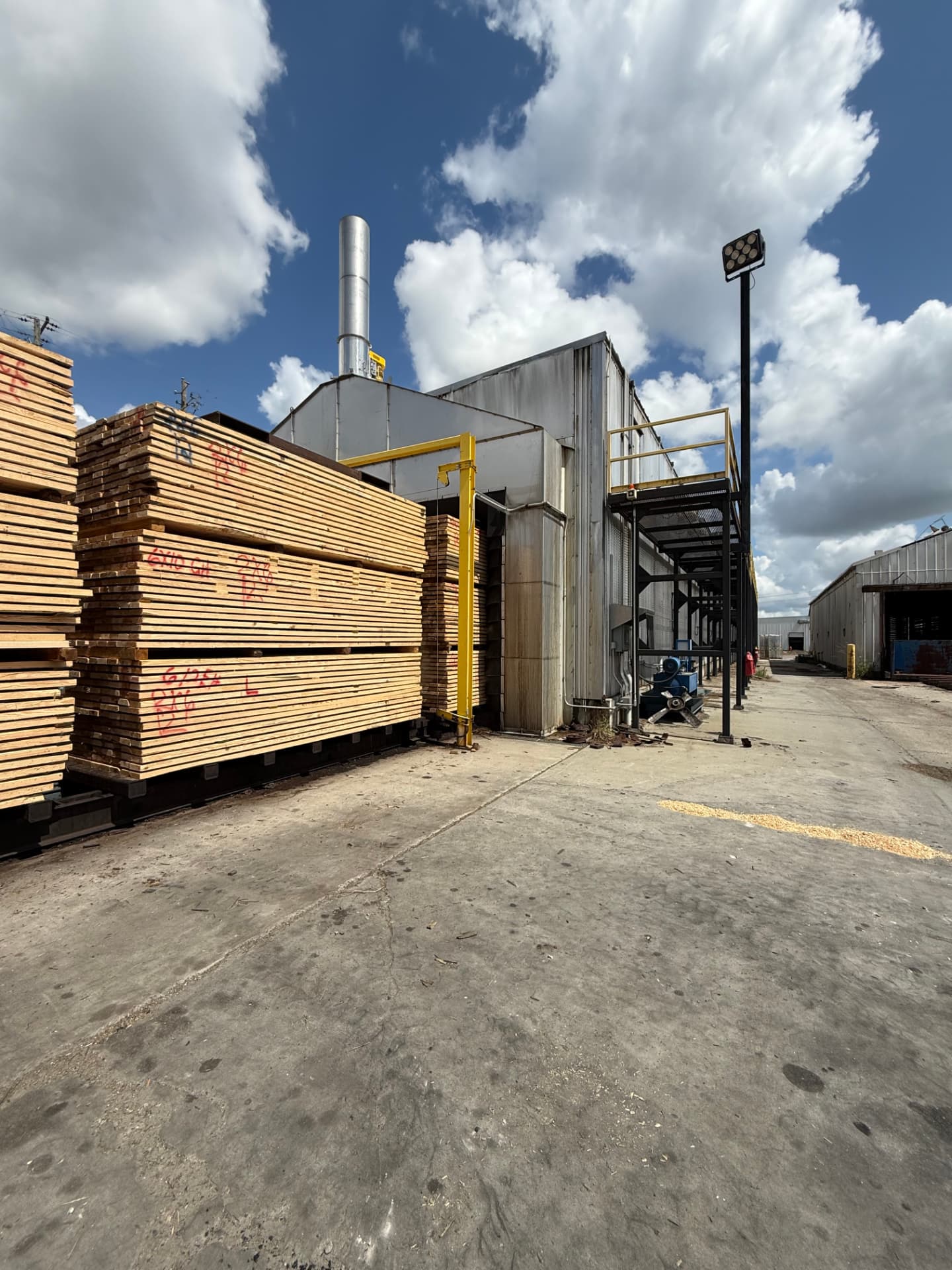 Lumber charge staged beside a dry kiln exterior with stack and blower at a mill